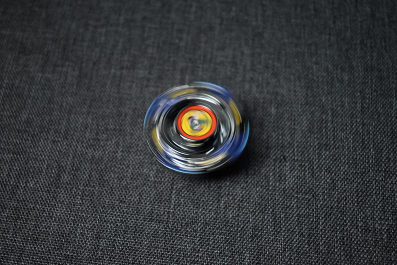 Op de afbeelding: Een draaiend speelgoed met een zwart, blauw en geel ontwerp. Het speelgoed heeft een transparante kunststof buitenlaag en een metalen binnenlaag.