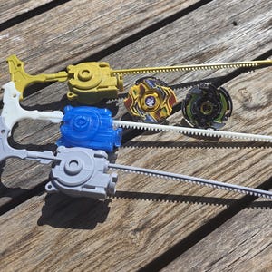 May include: A selection of spinning top toys, featuring yellow, blue, and grey launchers, and two spinning tops with detailed patterns. The toys are displayed on a weathered wooden surface.