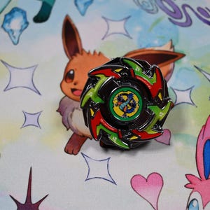 Op de afbeelding: Een zwarte, groene en rode spinning top pin met een centraal ontwerp. De pin is geplaatst tegen een kleurrijke achtergrond met stripfiguren en stervormen. De pin is rond met een gedetailleerd ontwerp.