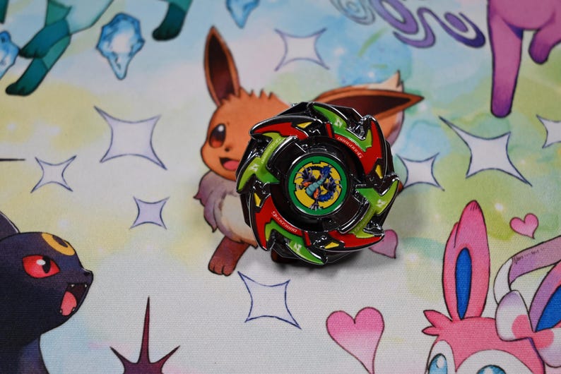 Op de afbeelding: Een zwarte, groene en rode spinning top pin met een centraal ontwerp. De pin is geplaatst tegen een kleurrijke achtergrond met stripfiguren en stervormen. De pin is rond met een gedetailleerd ontwerp.