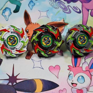 Op de afbeelding: Drie kleurrijke tolletjes met rode, groene en witte accenten. De tolletjes zijn geplaatst op een patroon met stripfiguren. Elk tolletje heeft een centraal embleem met een drakenontwerp.