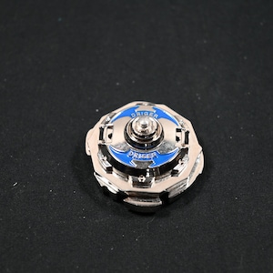 May include: A silver-colored spinning top toy with blue accents and the word "DRIGER" printed on it. The toy has a circular design with a central spinning mechanism. The background is a solid black color.