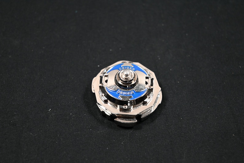 May include: A silver-colored spinning top toy with blue accents and the word "DRIGER" printed on it. The toy has a circular design with a central spinning mechanism. The background is a solid black color.