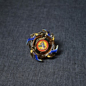Op de afbeelding: Een gouden en blauwe draaitop speelgoed met een rood, geel en blauw ontwerp. De top heeft een cirkelvormig midden met een afbeelding van een beest met vleugels.