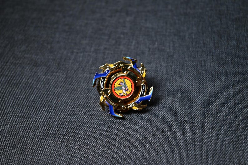 Op de afbeelding: Een gouden en blauwe draaitop speelgoed met een rood, geel en blauw ontwerp. De top heeft een cirkelvormig midden met een afbeelding van een beest met vleugels.