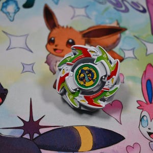 Op de afbeelding: Een witte, rode en groene tol met een centrale afbeelding van een draak. Het speelgoed staat op een kleurrijke achtergrond met stripfiguren. Het is een verzamelobject.