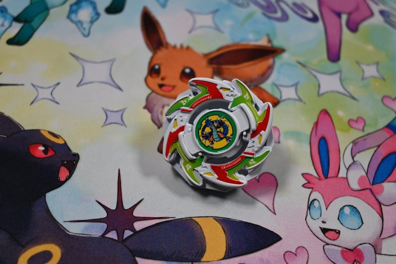 Op de afbeelding: Een witte, rode en groene tol met een centrale afbeelding van een draak. Het speelgoed staat op een kleurrijke achtergrond met stripfiguren. Het is een verzamelobject.