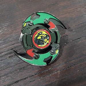 K&ouml;nnte beinhalten: Ein kreisf&ouml;rmiges Kreiselspielzeug mit einem gr&uuml;nen und schwarzen Rahmen mit roten Akzenten. Die Mitte enth&auml;lt ein kreisf&ouml;rmiges Emblem mit einer gelb-schwarzen Drachengrafik, umgeben von einem roten Rand mit japanischem Text in wei&szlig;en Zeichen.