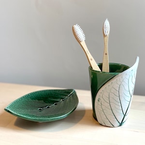 Puede incluir: Un jabonera de cerámica verde en forma de hoja y un portacepillos de dientes de cerámica verde con un diseño de hoja blanca. Dos cepillos de dientes de bambú con cerdas blancas están en el portacepillos.