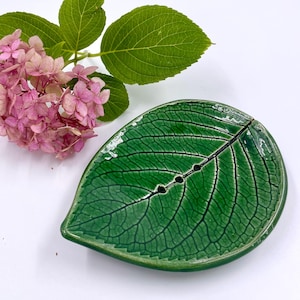 May include: A glossy green ceramic soap dish shaped like a leaf with a detailed vein texture and three small drainage holes along the center. Beside the dish is a cluster of pink hydrangea flowers with several bright green leaves.