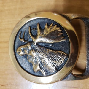 May include: A gold-toned belt buckle with a detailed moose head design. The moose is depicted in profile with its antlers raised. The buckle is circular and has a smooth, polished finish.