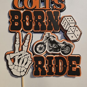 May include: A layered paper cake topper featuring the text COLT'S BORN TO RIDE in black and orange. The design includes a white skeleton hand making a peace sign, a black motorcycle illustration, and two white dice with black dots on a wooden stick.