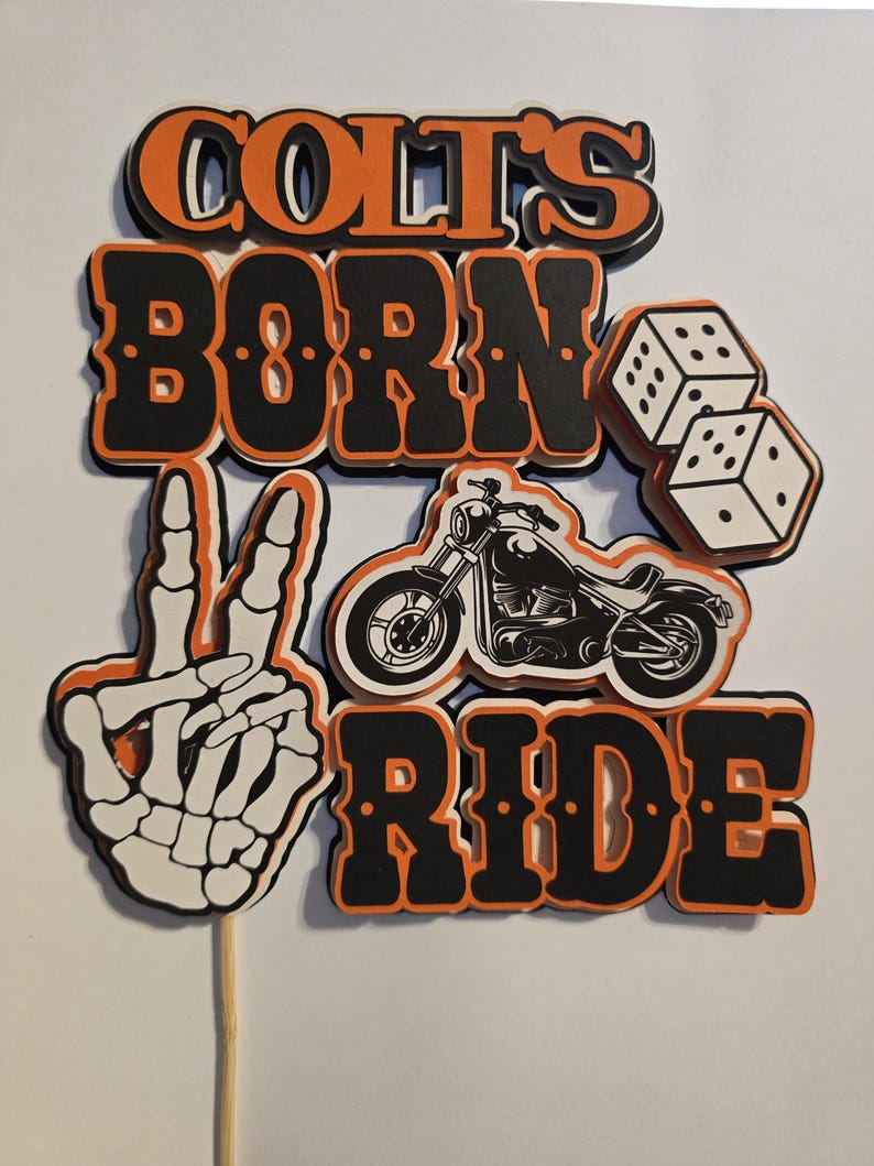 May include: A layered paper cake topper featuring the text COLT'S BORN TO RIDE in black and orange. The design includes a white skeleton hand making a peace sign, a black motorcycle illustration, and two white dice with black dots on a wooden stick.