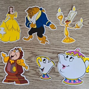 May include: A set of six paper cutouts featuring characters from the Disney movie Beauty and the Beast. The characters include Belle, Beast, Lumiere, Mrs. Potts, Chip, and Cogsworth. The cutouts are yellow, blue, red, and white.