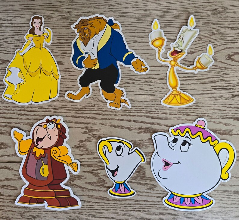 May include: A set of six paper cutouts featuring characters from the Disney movie Beauty and the Beast. The characters include Belle, Beast, Lumiere, Mrs. Potts, Chip, and Cogsworth. The cutouts are yellow, blue, red, and white.
