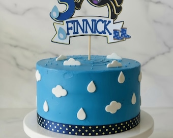 May include: A round blue cake decorated with white fondant clouds and raindrops, featuring a dark blue ribbon with yellow dots at the base. A cake topper displays the number 3, a cartoon spider, the name FINNICK, and blue rain boots.
