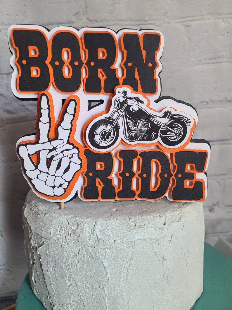 May include: A layered cake topper featuring the text BORN TO RIDE in black with orange outlines. The design includes a white skeleton hand making a peace sign and a black and white illustration of a motorcycle. The topper is mounted on a wooden stick.