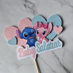 以下が含まれることがあります： リロ＆スティッチのキャラクター、スティッチとエンジェルのケーキトッパー。ピンクとブルーのハートのデザインで、「Baby Salazar」の文字が筆記体で書かれています。トッパーは木の棒に取り付けられています。