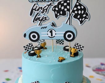 Puede incluir: Un pastel de dos niveles de color azul claro con una decoración de carreras que incluye un coche de carreras azul claro con el número uno, dos banderas a cuadros y el texto Connor's first lap, adornado con pequeños trofeos dorados y neumáticos.