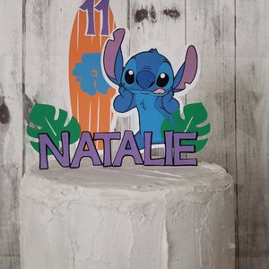 以下が含まれることがあります： 青い漫画のキャラクター、11の数字が入ったサーフボード、紫色の文字で「NATALIE」という名前がトッピングされた白いフロストケーキ。緑の熱帯の葉も含まれています。