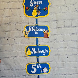 May include: A set of five blue and yellow signs with a Beauty and the Beast theme. The signs read "Be Our Guest", "Welcome to", "Aubrey's", "5th", and "Birthday". Each sign features a different character from the movie.