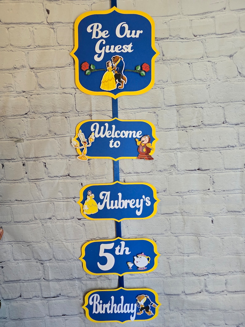 May include: A set of five blue and yellow signs with a Beauty and the Beast theme. The signs read "Be Our Guest", "Welcome to", "Aubrey's", "5th", and "Birthday". Each sign features a different character from the movie.