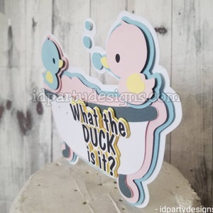 Puede incluir: Un adorno para pastel con dos patos de goma en una ba&ntilde;era con el texto "What the duck is it?" Los patos son azules y rosas con picos y patas amarillas. La ba&ntilde;era es blanca con detalles azules y rosas.