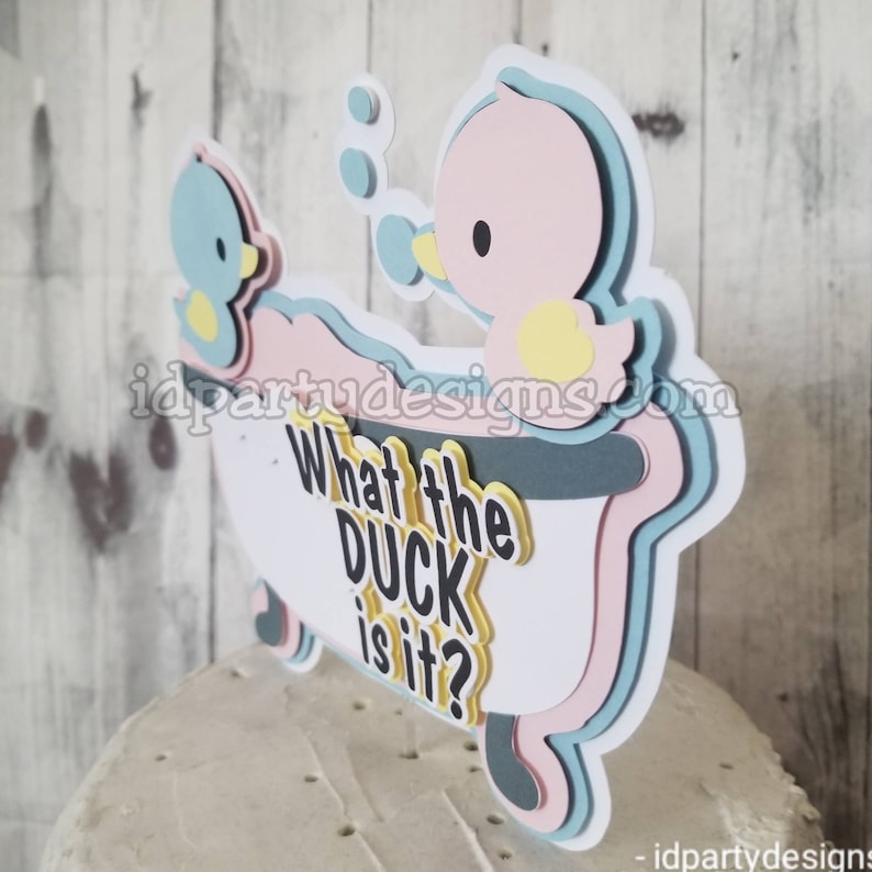 Puede incluir: Un adorno para pastel con dos patos de goma en una ba&ntilde;era con el texto "What the duck is it?" Los patos son azules y rosas con picos y patas amarillas. La ba&ntilde;era es blanca con detalles azules y rosas.