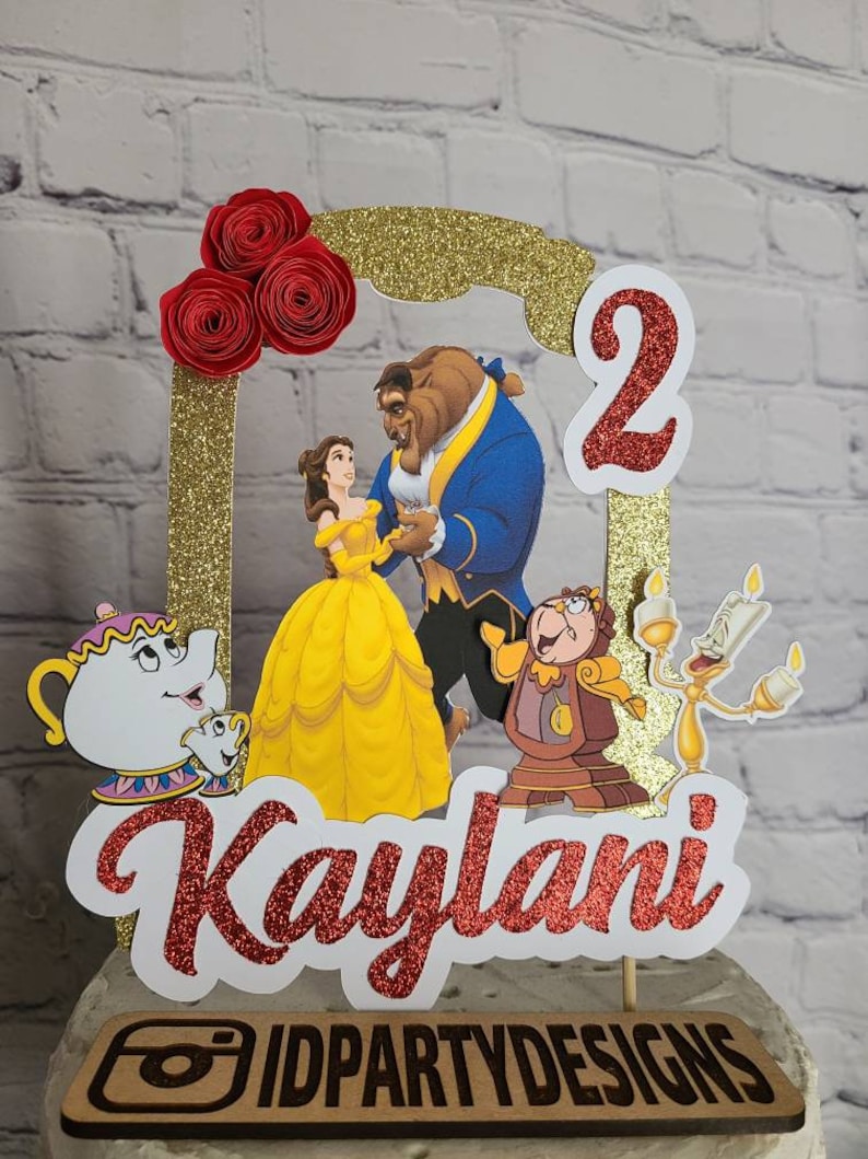 May include: A red and gold glitter cake topper with a picture of Belle and the Beast from Beauty and the Beast. The topper also includes the number 2, the name Kaylani, and the logo IDPARTYDESIGNS.