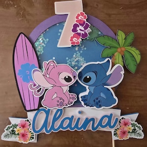 以下が含まれることがあります： スティッチとエンジェルのキャラクター、サーフボード、熱帯の花をあしらったカラフルなケーキトッパー。キャラクターの上には数字の7が表示され、下には青いスクリプトで「Alaina」という名前が書かれています。背景はグリッターが入った青い円です。