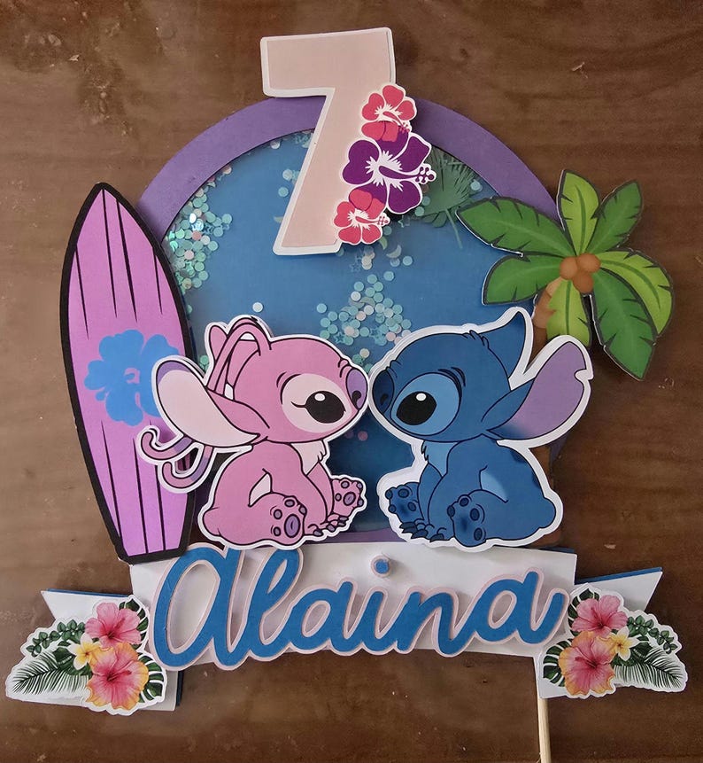 以下が含まれることがあります： スティッチとエンジェルのキャラクター、サーフボード、熱帯の花をあしらったカラフルなケーキトッパー。キャラクターの上には数字の7が表示され、下には青いスクリプトで「Alaina」という名前が書かれています。背景はグリッターが入った青い円です。