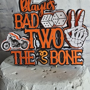 May include: A multi-layered cake topper featuring the orange and black text "Blayke's BAD TO THE BONE" with a "TWO" in the center. The design includes a black motorcycle, two white dice, and a white skeleton hand making a peace sign gesture.