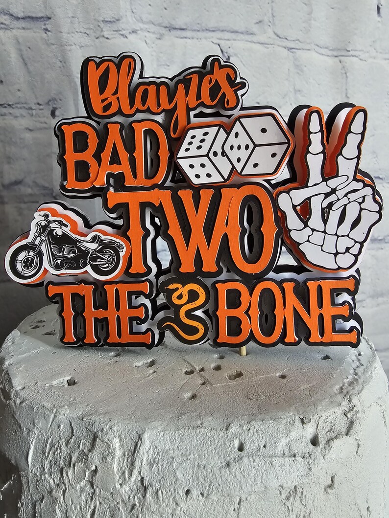May include: A multi-layered cake topper featuring the orange and black text "Blayke's BAD TO THE BONE" with a "TWO" in the center. The design includes a black motorcycle, two white dice, and a white skeleton hand making a peace sign gesture.