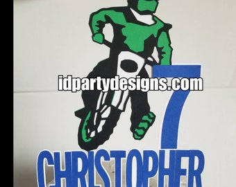 May include: A layered paper cake topper featuring a green and black motocross rider on a motorcycle, a large blue number 7, and the name CHRISTOPHER in blue letters. The text idpartydesigns.com is printed across the center of the design.