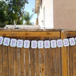 以下が含まれることがあります： 「HAPPY BIRTHDAY」の文字がラベンダー色の文字で、水色の切り抜きに書かれたお祝いのバナー。ピンク色の紐で吊るされており、両端には漫画のキャラクターの画像が描かれています。木製のフェンスに吊るされています。