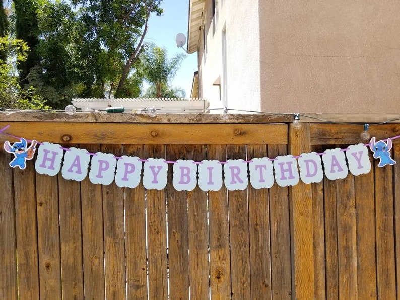 以下が含まれることがあります： 「HAPPY BIRTHDAY」の文字がラベンダー色の文字で、水色の切り抜きに書かれたお祝いのバナー。ピンク色の紐で吊るされており、両端には漫画のキャラクターの画像が描かれています。木製のフェンスに吊るされています。