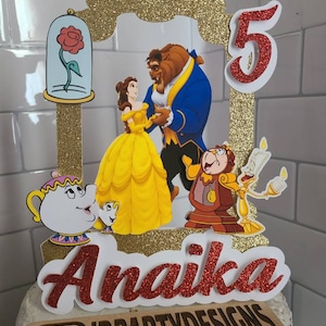 May include: A red glitter cake topper with a gold glitter frame featuring Belle and the Beast from Beauty and the Beast. The topper also includes a red glitter number 5, a red glitter name "Anaika", and a wooden sign with the text "IDPARTYDESIGNS".