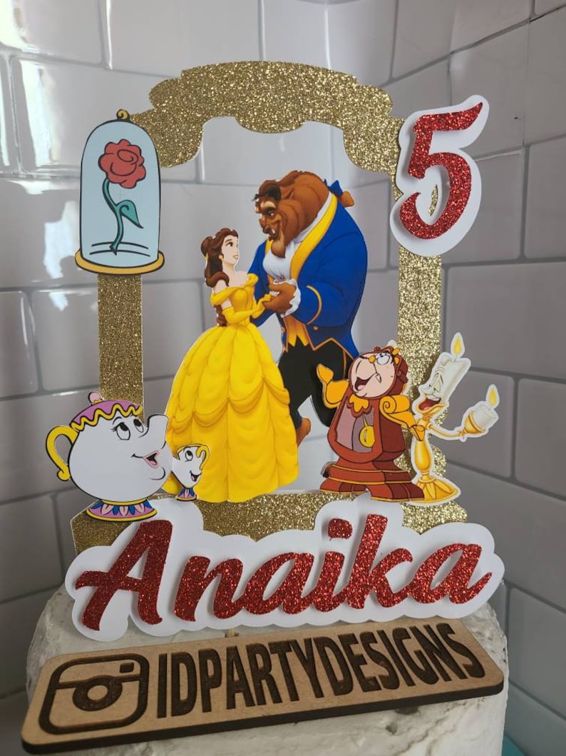 May include: A red glitter cake topper with a gold glitter frame featuring Belle and the Beast from Beauty and the Beast. The topper also includes a red glitter number 5, a red glitter name "Anaika", and a wooden sign with the text "IDPARTYDESIGNS".