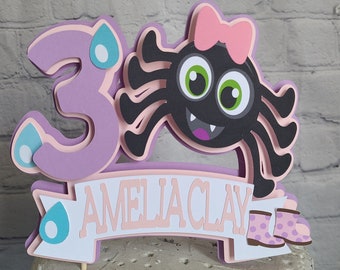 May include: Purple and pink cake topper with a black spider wearing a pink bow, the number 3, and the name "Amelia Clay".