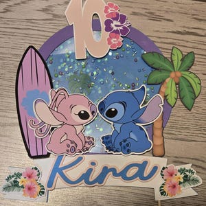 以下が含まれることがあります： スティッチとエンジェルのキャラクター、サーフボード、ヤシの木をあしらったケーキトッパー。キャラクターの上には数字の10、下には「Kira」という名前が表示されています。背景は青色で、キラキラしています。