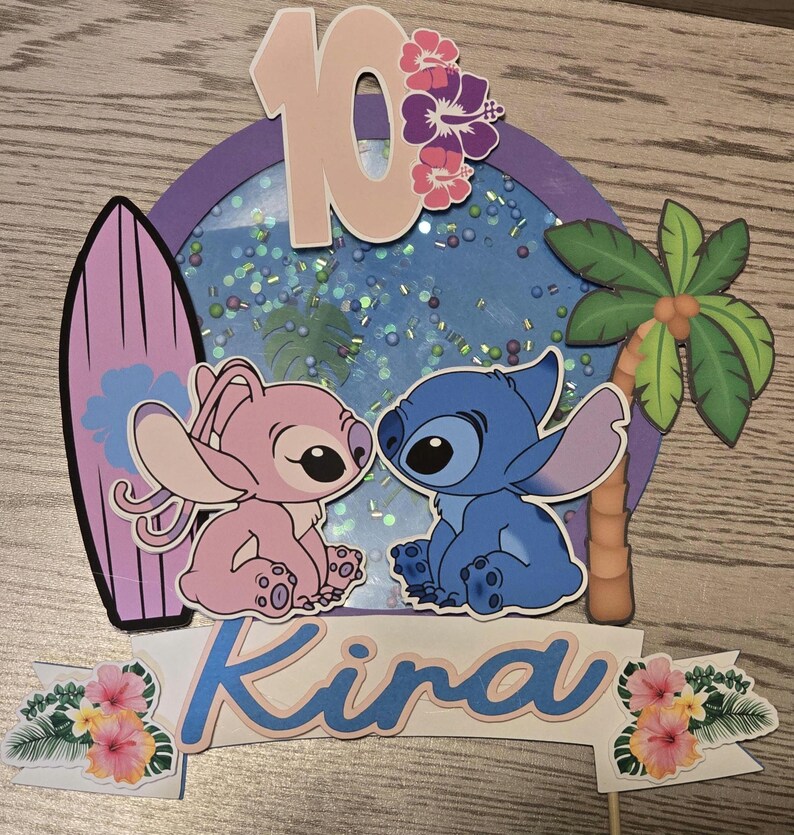 以下が含まれることがあります： スティッチとエンジェルのキャラクター、サーフボード、ヤシの木をあしらったケーキトッパー。キャラクターの上には数字の10、下には「Kira」という名前が表示されています。背景は青色で、キラキラしています。