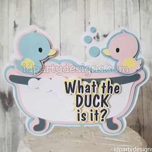 Puede incluir: Un adorno para pastel de pato azul y rosa con el texto "What the DUCK is it?" en letras negras. Los patos est&aacute;n en una ba&ntilde;era con espuma blanca y burbujas azules.