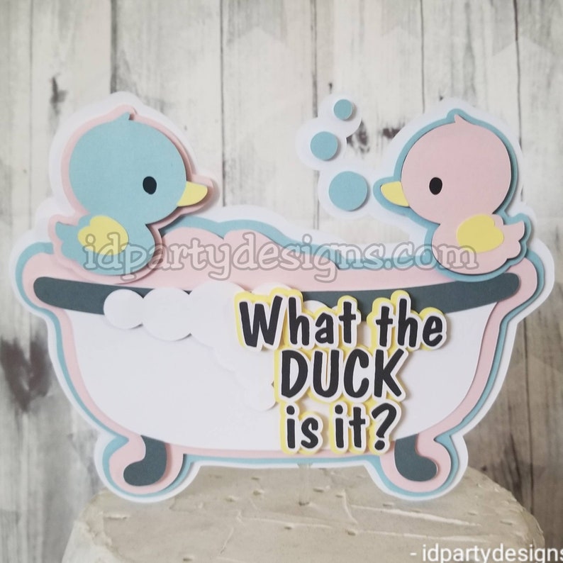 Puede incluir: Un adorno para pastel de pato azul y rosa con el texto "What the DUCK is it?" en letras negras. Los patos est&aacute;n en una ba&ntilde;era con espuma blanca y burbujas azules.