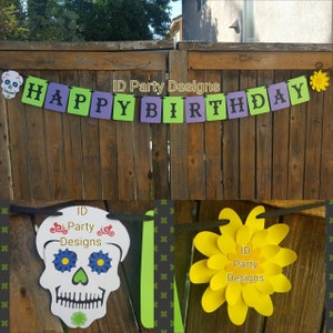 May include: A festive "Happy Birthday" banner with green and purple lettered flags. The banner is decorated with a sugar skull and a yellow paper flower. The banner is strung across a wooden fence.