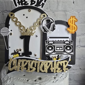 May include: A black and gold cake topper with the words "The Big 1 Christopher" and decorations including a microphone, headphones, a gold chain, a crown, a boombox, and a dollar sign.