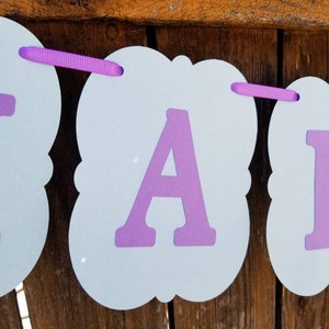 以下が含まれることがあります： 紫色の文字と紫色のリボンが付いた水色のバナー。バナーは紙製で、装飾的な形をしています。背景は木製の表面です。