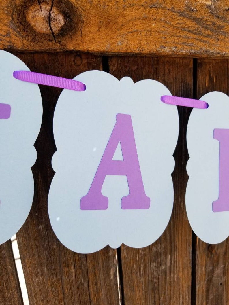 以下が含まれることがあります： 紫色の文字と紫色のリボンが付いた水色のバナー。バナーは紙製で、装飾的な形をしています。背景は木製の表面です。