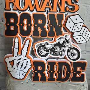 May include: A layered cake topper featuring the text ROWAN'S BORN TO RIDE in black and orange lettering. The design includes a white skeleton hand making a peace sign, a black motorcycle illustration, and two white dice with black dots.
