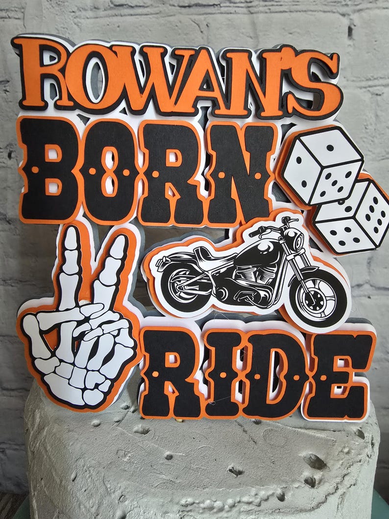 May include: A layered cake topper featuring the text ROWAN'S BORN TO RIDE in black and orange lettering. The design includes a white skeleton hand making a peace sign, a black motorcycle illustration, and two white dice with black dots.