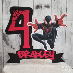 May include: Red and black cake topper featuring a cartoon Spider-Man character with a web design and the number 4. A black banner with the name Bradley in red letters is below the character.